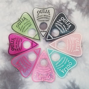 May include: Six different colored Ouija boards in the shape of a triangle. Each board has a different color background, including pink, green, black, white, and two shades of pink. The boards have a white outline and black lettering. The boards are arranged in a circle.