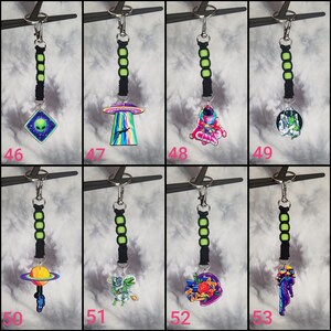 May include: Eight colorful keychains with different space-themed designs. Each keychain features a black and green cord with a silver ring. The designs include an alien, a UFO, an astronaut, a space creature, a planet, a cat, a flower, and a mermaid.