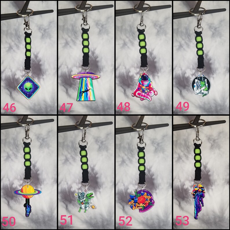 May include: Eight colorful keychains with different space-themed designs. Each keychain features a black and green cord with a silver ring. The designs include an alien, a UFO, an astronaut, a space creature, a planet, a cat, a flower, and a mermaid.