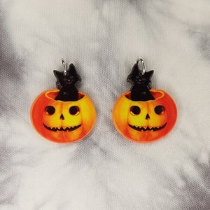 May include: A pair of orange and yellow pumpkin earrings with black cats peeking out of the top. The earrings are made of acrylic and have silver hooks.