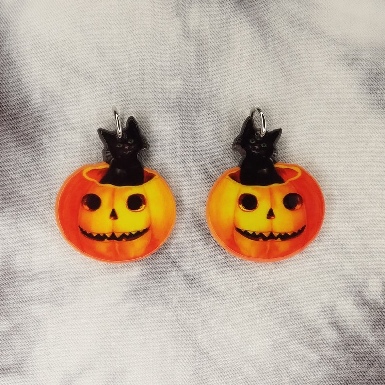 May include: A pair of orange and yellow pumpkin earrings with black cats peeking out of the top. The earrings are made of acrylic and have silver hooks.