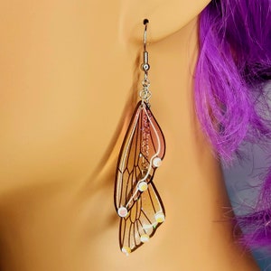 Puede incluir: Un par de pendientes colgantes con un dise&ntilde;o de mariposa en capas con alas iridiscentes. Las alas son transl&uacute;cidas y tienen una combinaci&oacute;n de colores rosa y marr&oacute;n con detalles blancos.