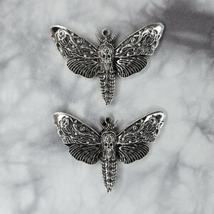 Puede incluir: Dos abalorios de metal plateado en forma de polilla con detalles intrincados. Las polillas tienen un dise&ntilde;o de calavera en sus cuerpos y son perfectas para la elaboraci&oacute;n de joyas o manualidades.