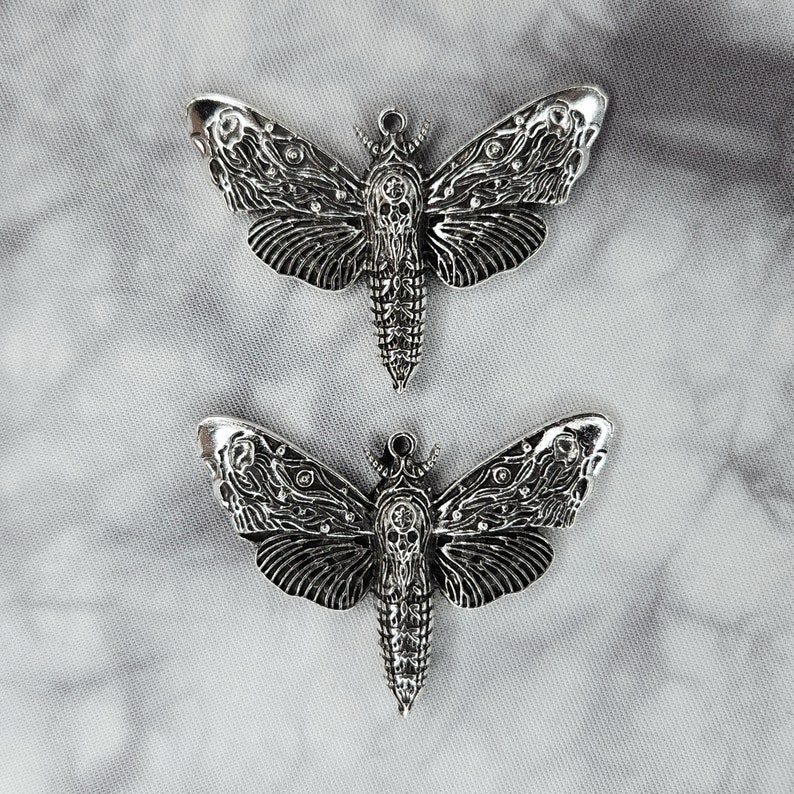 Puede incluir: Dos abalorios de metal plateado en forma de polilla con detalles intrincados. Las polillas tienen un dise&ntilde;o de calavera en sus cuerpos y son perfectas para la elaboraci&oacute;n de joyas o manualidades.