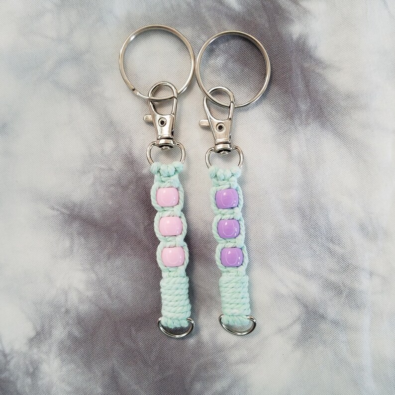 May include: Two keychains with light green string and colorful beads. One keychain has pink beads and the other has purple beads.