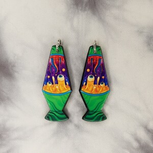 May include: A pair of green and yellow lava lamp earrings with a psychedelic design. The earrings feature a swirling pattern of purple, blue, and yellow, with two large eyes in the center.