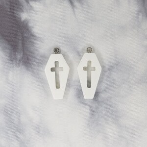 May include: A pair of white coffin-shaped earrings with a cross cut-out. The earrings have silver jump rings.