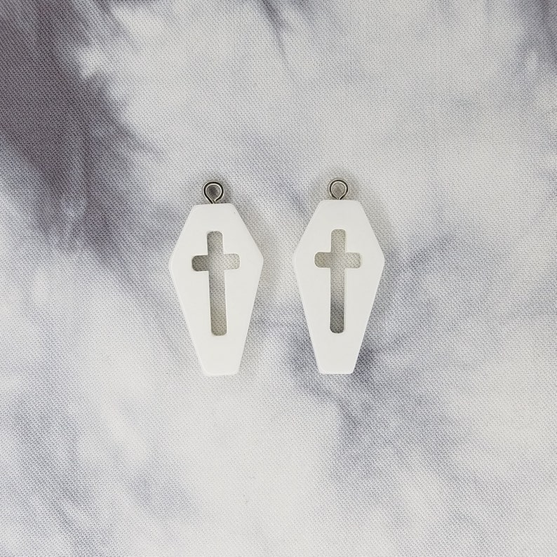 May include: A pair of white coffin-shaped earrings with a cross cut-out. The earrings have silver jump rings.