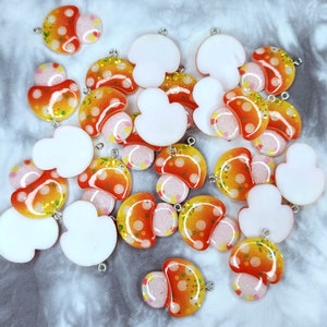May include: A collection of mushroom-shaped charms in various stages of completion. The charms feature a mix of orange, yellow, and white colors with glitter accents and polka dots. Each charm has a small silver loop for attaching to jewelry or other crafts.