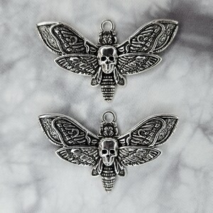Puede incluir: Dos abalorios de metal plateados en forma de esfinge de la muerte. Las polillas tienen detalles intrincados y una cabeza en forma de calavera.