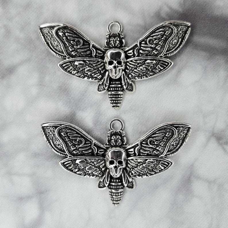 Puede incluir: Dos abalorios de metal plateados en forma de esfinge de la muerte. Las polillas tienen detalles intrincados y una cabeza en forma de calavera.