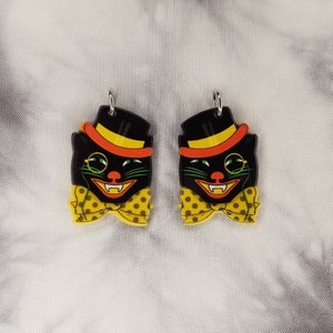 May include: A pair of black cat earrings with yellow and orange accents. The cats are wearing top hats and bow ties with white polka dots.