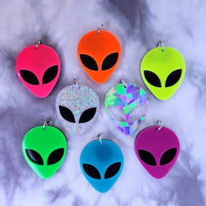 May include: Set of ten alien-shaped acrylic charms in various colors, including green, blue, pink, orange, silver, and holographic. Each charm features a black alien face with large eyes.