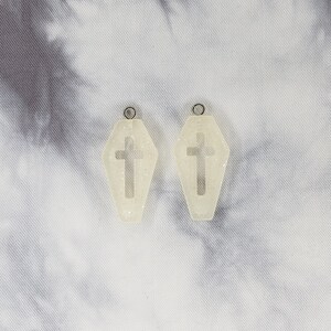 May include: A pair of white coffin-shaped earrings with a cross cutout. The earrings are made of resin and have a glittery finish.