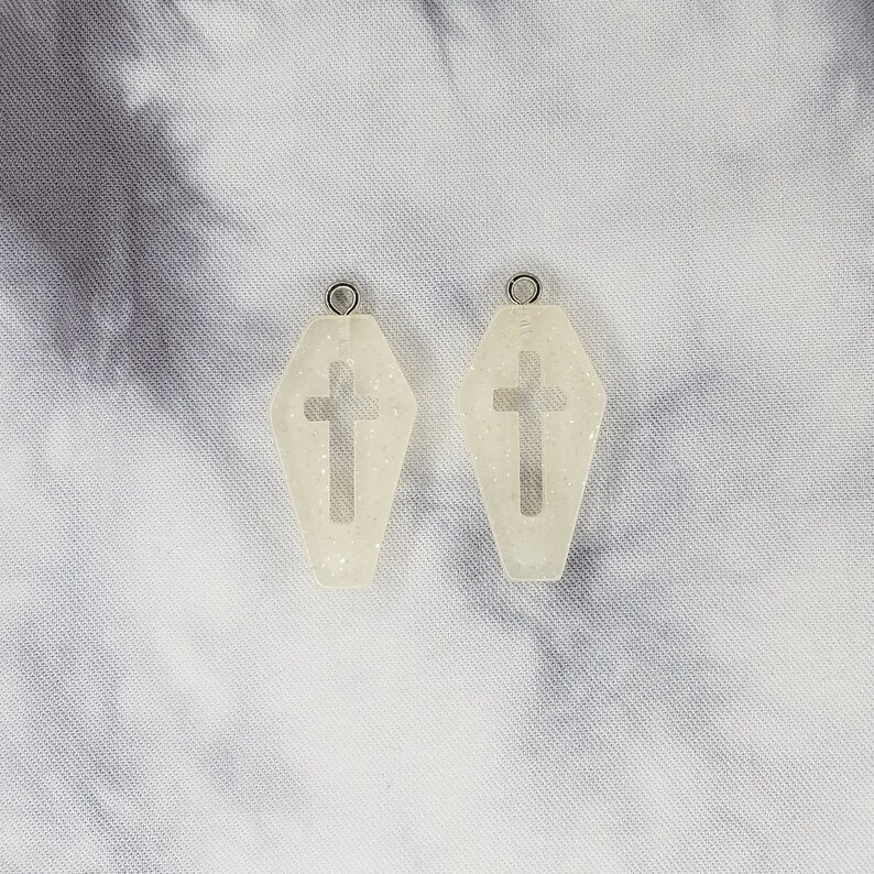 May include: A pair of white coffin-shaped earrings with a cross cutout. The earrings are made of resin and have a glittery finish.