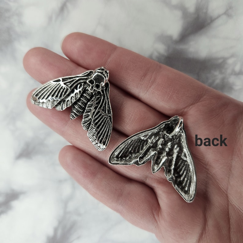 Puede incluir: Dos pendientes de plata en forma de polilla, uno con las alas extendidas y el otro con las alas cerradas. La parte posterior del pendiente es visible en la imagen.