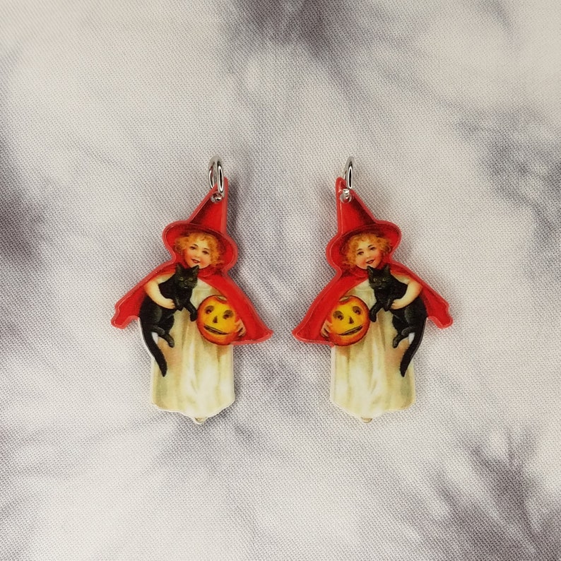 May include: A pair of Halloween-themed earrings featuring a vintage illustration of a girl dressed as a witch holding a black cat and a jack-o'-lantern.