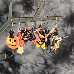 Pode incluir: Um conjunto de seis brincos temáticos de Halloween com gatos pretos, abóboras e chapéus de bruxa. Os brincos são feitos de acrílico colorido e têm ganchos prateados.