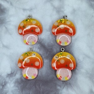 May include: Four mushroom-shaped charms with a silver loop at the top. The caps are orange and yellow with white polka dots and glitter. The stems are white with colorful confetti.