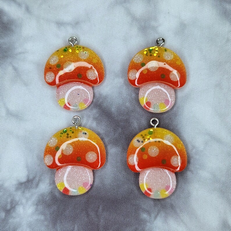 May include: Four mushroom-shaped charms with a silver loop at the top. The caps are orange and yellow with white polka dots and glitter. The stems are white with colorful confetti.