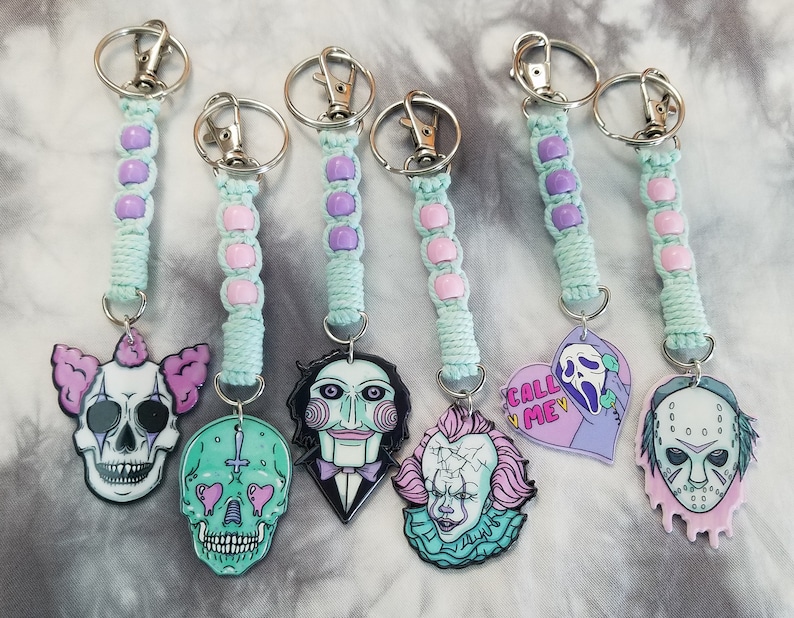 May include: Six keychains with pastel green and purple beaded strings and various horror movie character charms. The charms include a clown skull, a skull with hearts for eyes, a character with a red and black mask, a clown with a cracked face, a heart with the text "Call Me" and a ghost face, and a hockey mask with dripping blood.