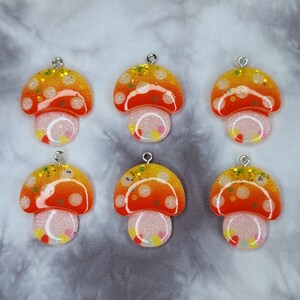 May include: Six mushroom-shaped pendants with silver-colored loops. The pendants have orange and yellow caps with white polka dots and glitter. The stems are white with colorful confetti-like accents. The background is a gray and white tie-dye pattern.
