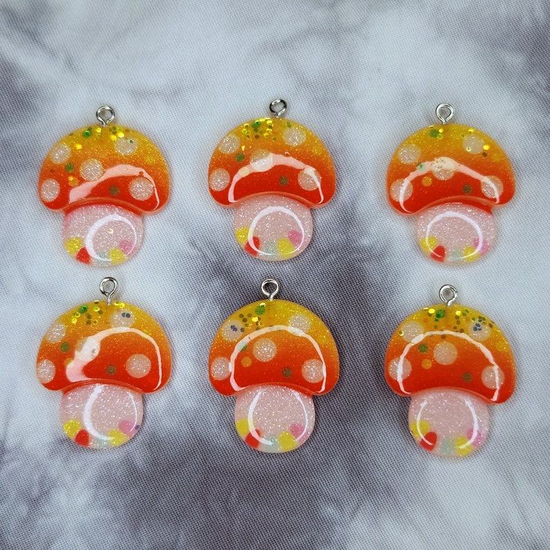 May include: Six mushroom-shaped pendants with silver-colored loops. The pendants have orange and yellow caps with white polka dots and glitter. The stems are white with colorful confetti-like accents. The background is a gray and white tie-dye pattern.