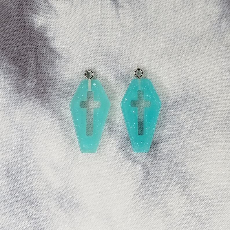 May include: A pair of turquoise blue coffin-shaped earrings with a cross cutout. The earrings have a glitter finish and silver jump rings.