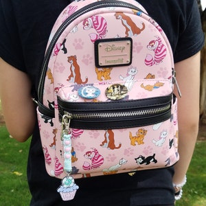 May include: A pink Disney Loungefly mini backpack featuring a repeating pattern of cats from the animated film The Aristocats. The backpack has a black zipper and a black adjustable strap. The backpack has a charm attached to the zipper pull. The charm is a cupcake with a green frosting swirl and a pink cherry on top.
