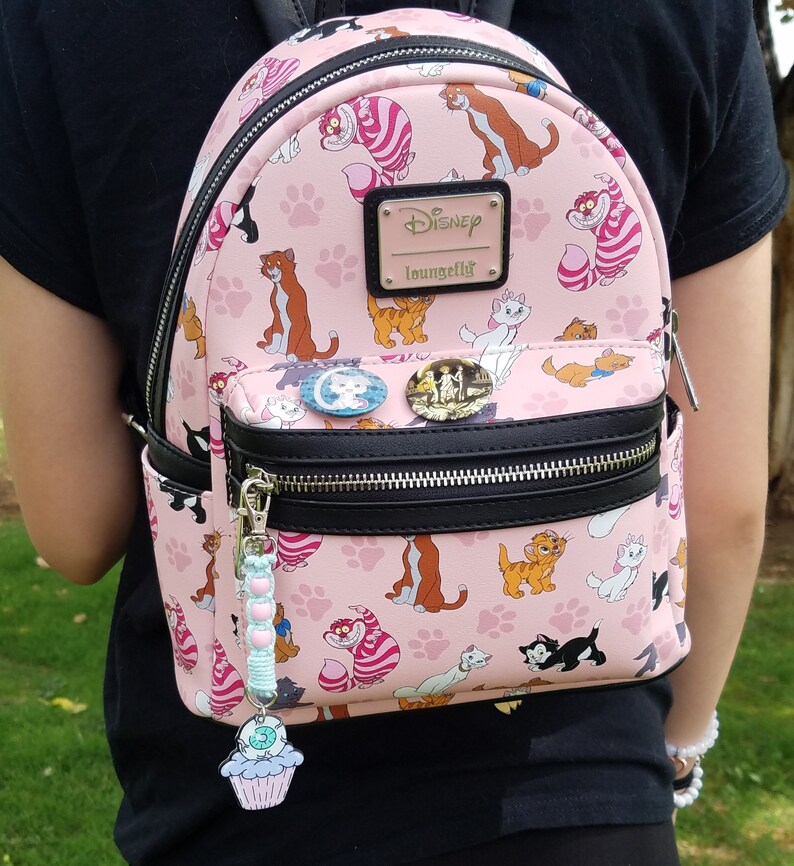 May include: A pink Disney Loungefly mini backpack featuring a repeating pattern of cats from the animated film The Aristocats. The backpack has a black zipper and a black adjustable strap. The backpack has a charm attached to the zipper pull. The charm is a cupcake with a green frosting swirl and a pink cherry on top.