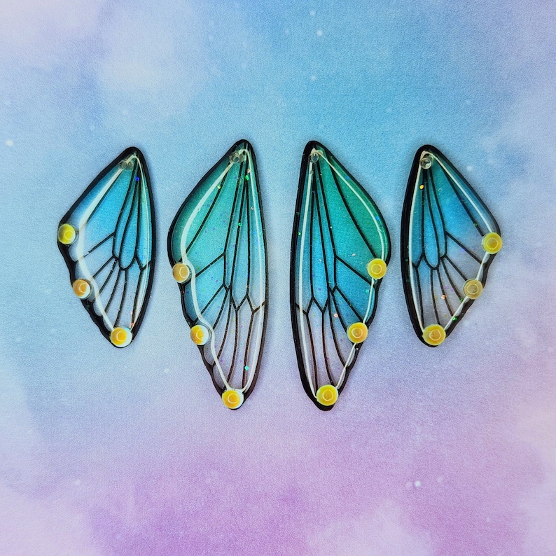 Puede incluir: Cuatro abalorios de acr&iacute;lico en forma de ala de mariposa azul y verde iridiscente con detalles amarillos.