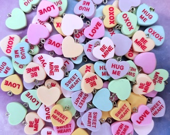 May include: A collection of colorful heart-shaped candy charms with various messages, including "Hug Me", "Be Mine", "Sweet Heart", "XOXO", and "Love".