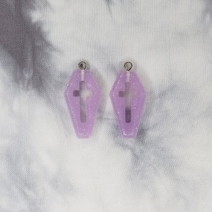 May include: A pair of purple glitter coffin-shaped earrings with a cross cutout. The earrings have silver jump rings.