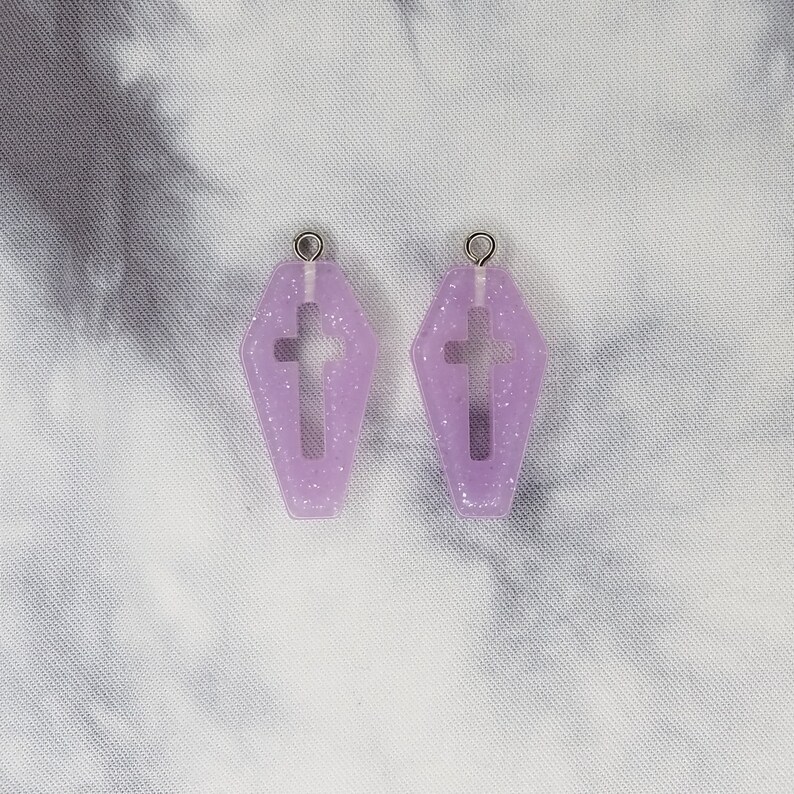 May include: A pair of purple glitter coffin-shaped earrings with a cross cutout. The earrings have silver jump rings.