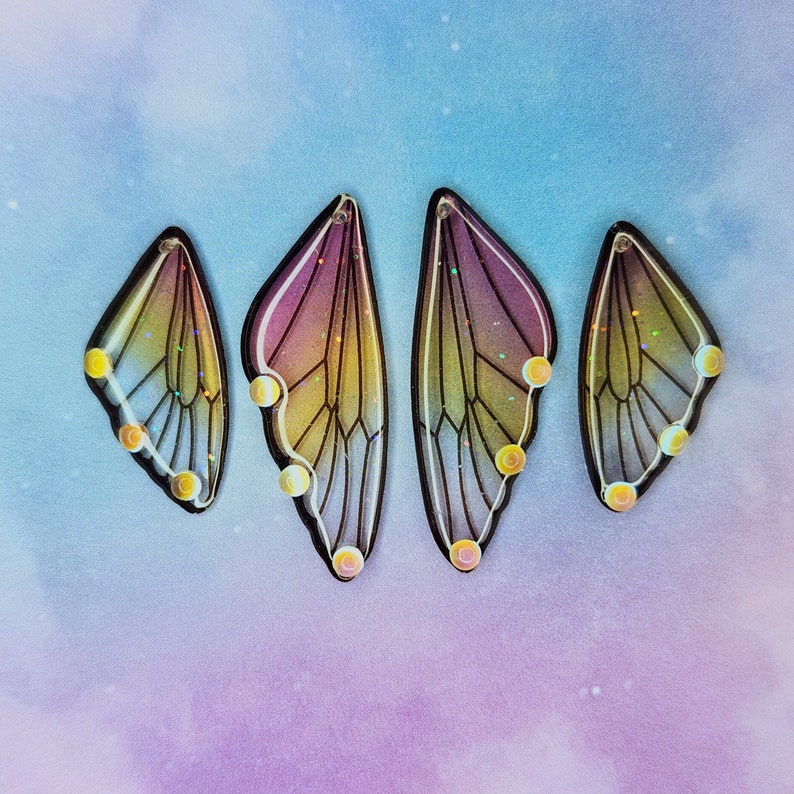 Puede incluir: Cuatro encantos iridiscentes en forma de ala de mariposa con un degradado rosa y amarillo y detalles de purpurina dorada.