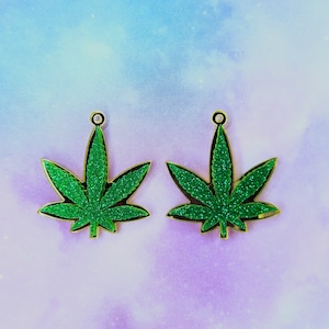 May include: Two green glitter marijuana leaf earrings with gold-colored metal earring hooks.