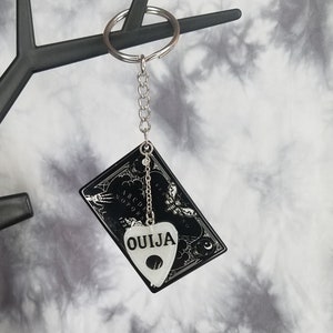 以下が含まれることがあります： 銀色のチェーンと銀色のプランシェットチャームが付いた、白黒のウィジャボードキーチェーン。プランシェットチャームはハート型で、「OUIJA」の文字が書かれています。