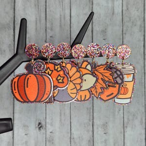 Puede incluir: Conjunto de seis pendientes de temática otoñal con detalles de purpurina. Los pendientes presentan una calabaza, un pavo, un zorro, una hoja, una taza de café y un bastón de caramelo.
