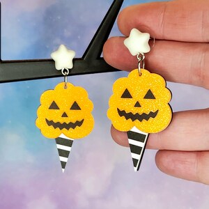 K&ouml;nnte beinhalten: Ein Paar Halloween-Ohrringe mit einem gelben und orangefarbenen K&uuml;rbis-f&ouml;rmigen Zuckerwatte mit einem schwarz-wei&szlig; gestreiften Kegel. Jeder Ohrring hat oben einen wei&szlig;en Stern-Anh&auml;nger.
