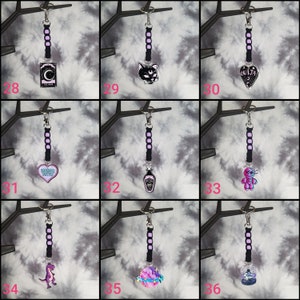 May include: Nine black keychains with different charms. The charms include a crescent moon, a black cat, a heart with the text "Greatest Love Story", a potion bottle, a voodoo doll, a dinosaur, a planet, a skull, and a heart with the text "Ouija".