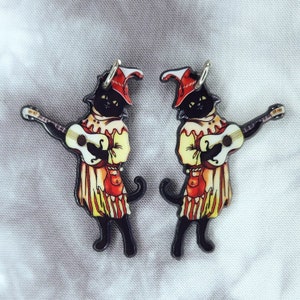 May include: A pair of black cat earrings with a jester hat and playing a guitar. The cats are wearing red and yellow clothing.