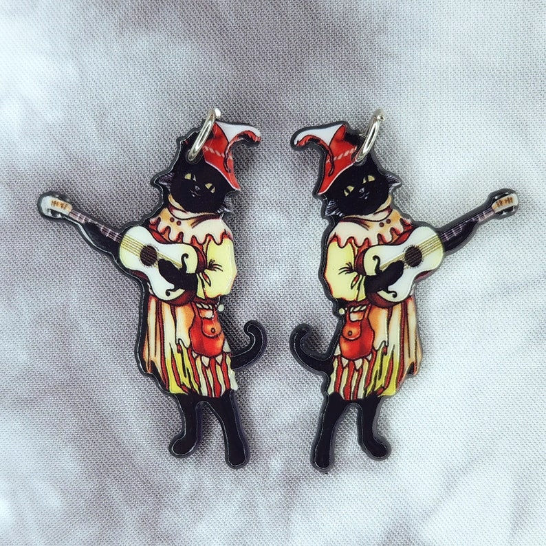 May include: A pair of black cat earrings with a jester hat and playing a guitar. The cats are wearing red and yellow clothing.
