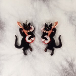 May include: A pair of black cat earrings with a banjo. The cats are wearing a white ruffle collar and are playing the banjo with their front paws. The banjo is red, white, and blue.