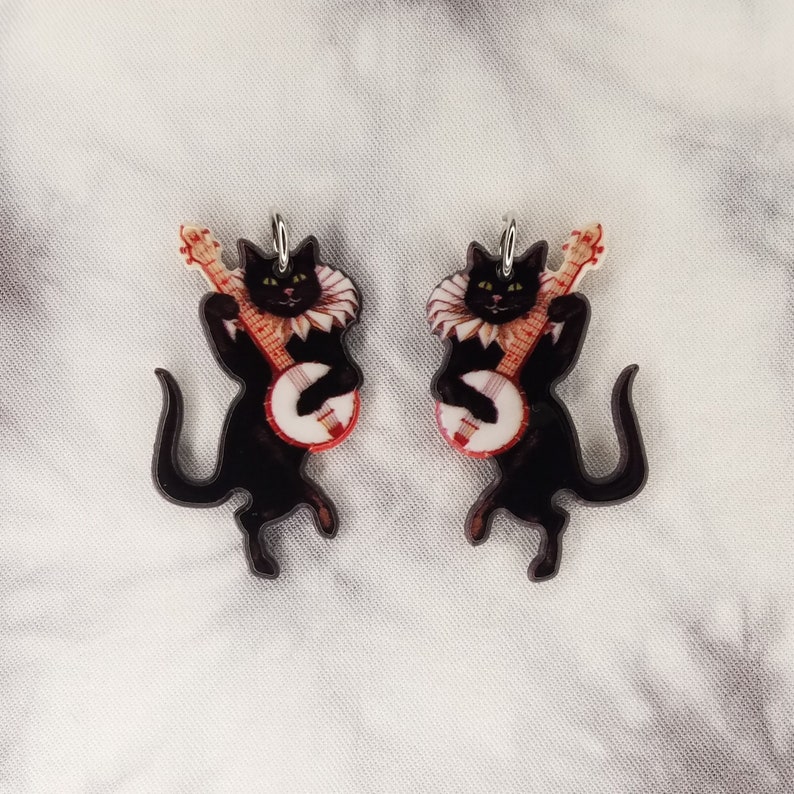 May include: A pair of black cat earrings with a banjo. The cats are wearing a white ruffle collar and are playing the banjo with their front paws. The banjo is red, white, and blue.