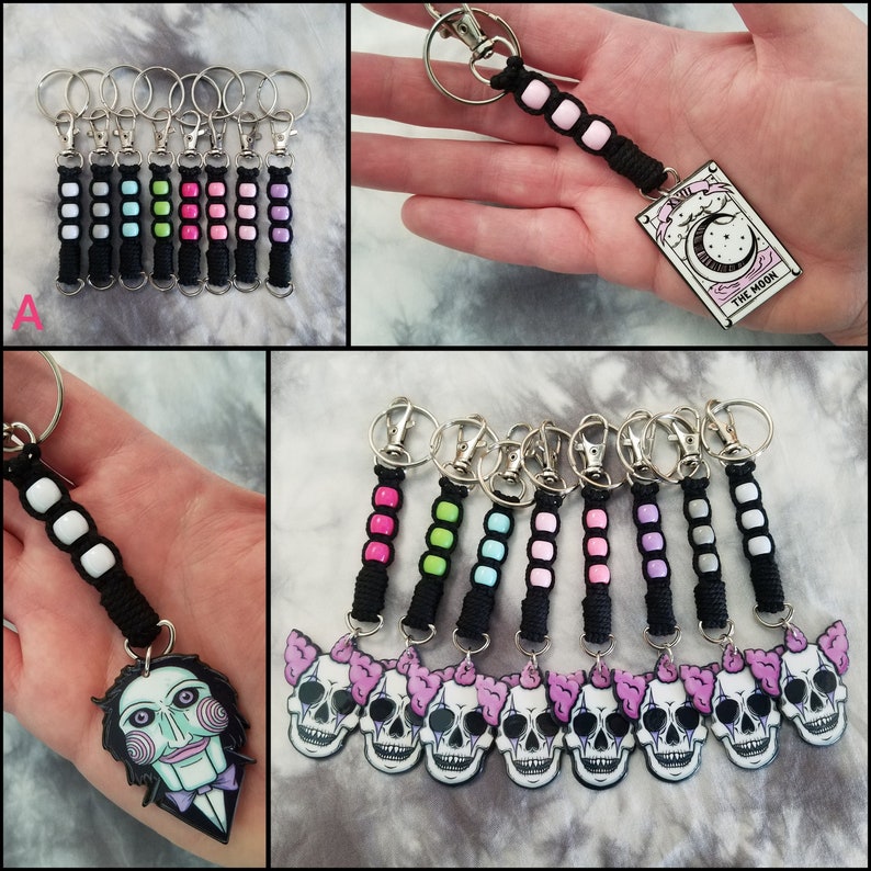 May include: A set of 10 colorful keychains with different charms. The charms include a skull with pink and white details, a puppet with a red mouth and black eyes, and a tarot card with the text "The Moon".