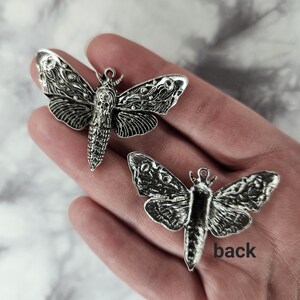 Puede incluir: Dos abalorios de polilla plateados, uno con las alas extendidas y el otro con las alas cerradas, con la palabra "back" escrita en la mano que sostiene el abalorio con las alas cerradas.
