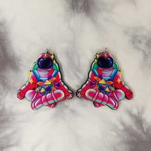 May include: A pair of pink astronaut earrings with a mushroom design. The astronauts are in a meditative pose with their eyes closed. The mushrooms are colorful and have a psychedelic vibe.