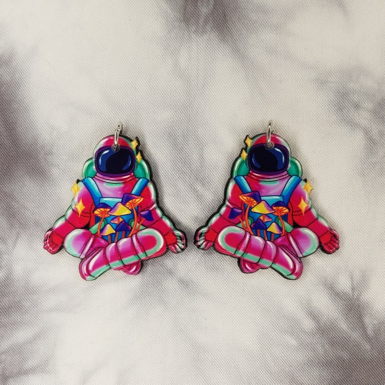 May include: A pair of pink astronaut earrings with a mushroom design. The astronauts are in a meditative pose with their eyes closed. The mushrooms are colorful and have a psychedelic vibe.