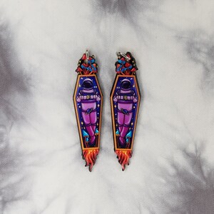 May include: A pair of astronaut-themed earrings with a purple astronaut in a coffin with a rocket-shaped flame. The astronaut is surrounded by mushrooms and stars.