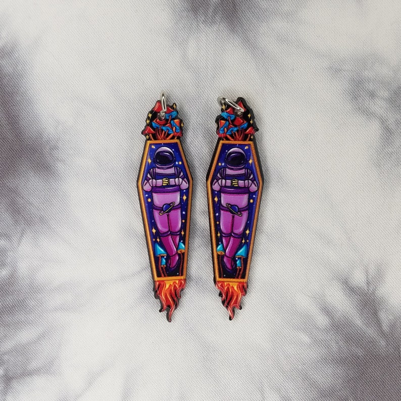 May include: A pair of astronaut-themed earrings with a purple astronaut in a coffin with a rocket-shaped flame. The astronaut is surrounded by mushrooms and stars.